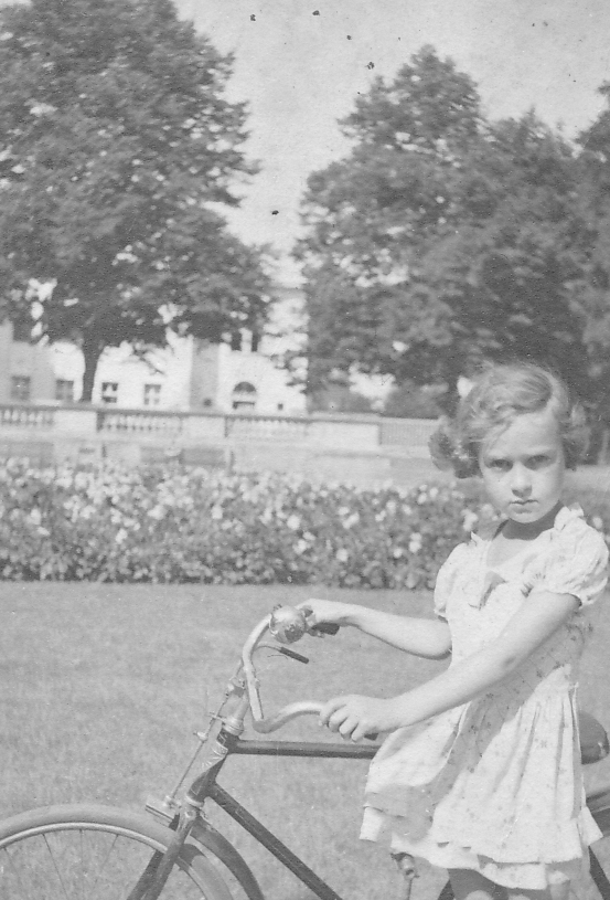 Rita Thomas alias Tommy als Kind im Kleid auf einem Fahrrad; im Hintergrund parkähnliche Anlage vor Gebäude