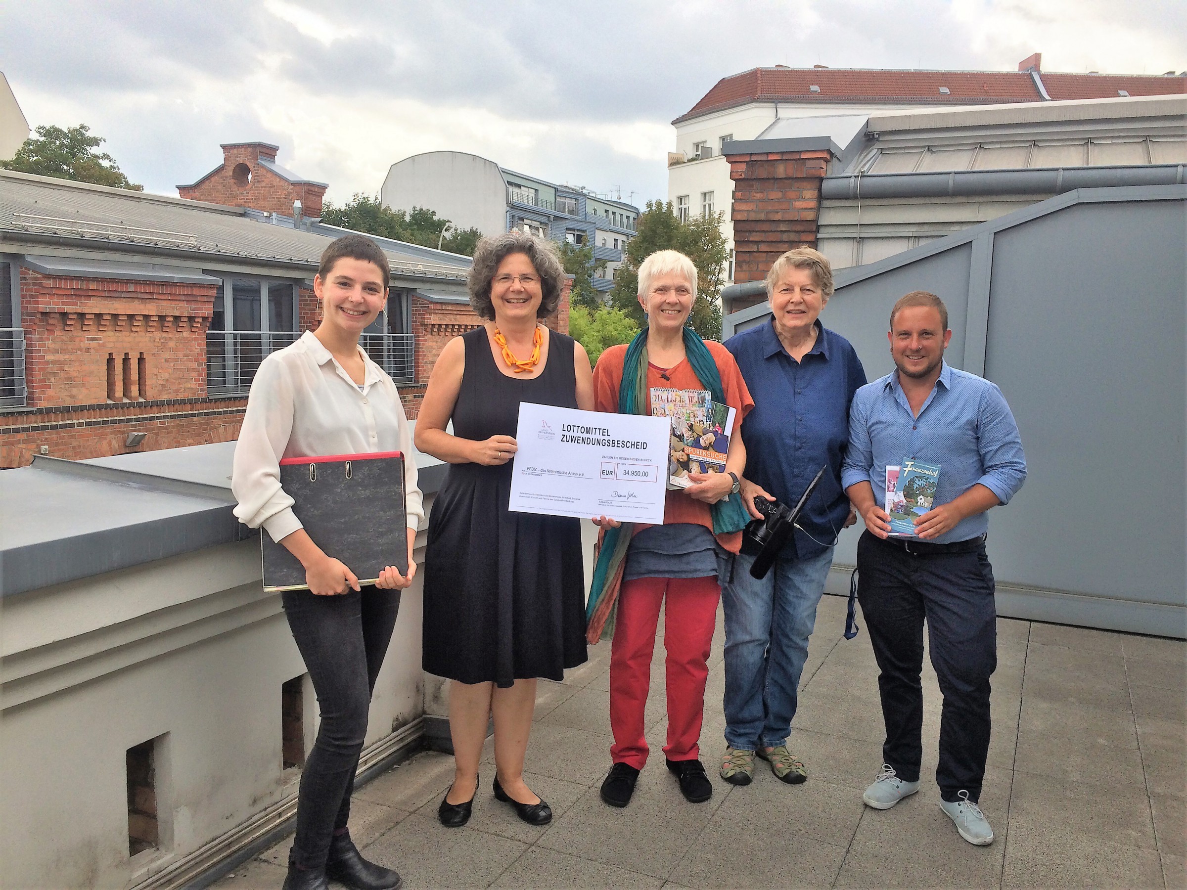 Fünf Personen auf der Dachterrasse (v.l.n.r.): FFBIZ-Mitarbeiterin Lisa Schug, Almuth Hartwig-Tiedt, Tina Kuhne von der Kontakt- und Koordinierungsstelle für Mädchenarbeit im Land Brandenburg (KuKMA), Tille Ganz, FFBIZ-Projektleiter Roman Klarfeld; Almuth Hartwig-Tiedt hält ein Schild hoch mit der Bezeichnung Lottomittel Zuwendungsbescheid