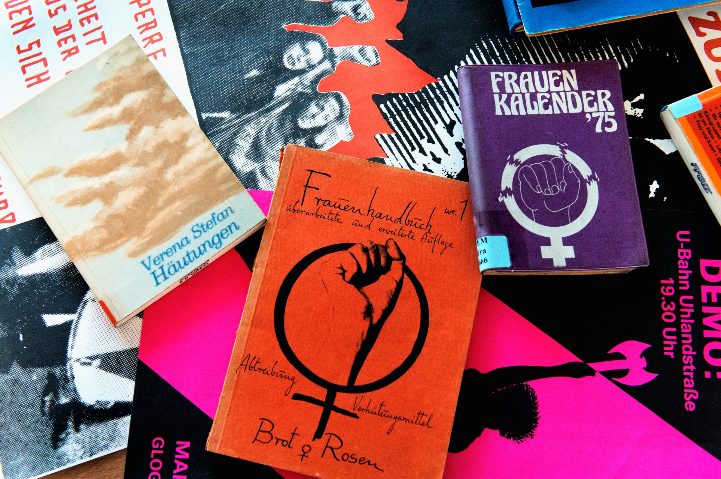 diverse Publikationen und Plakate neben und übereinander angeordnet u.a. das rote Cover des Frauenhandbuches und das lilafarbene Cover des Frauenkalenders