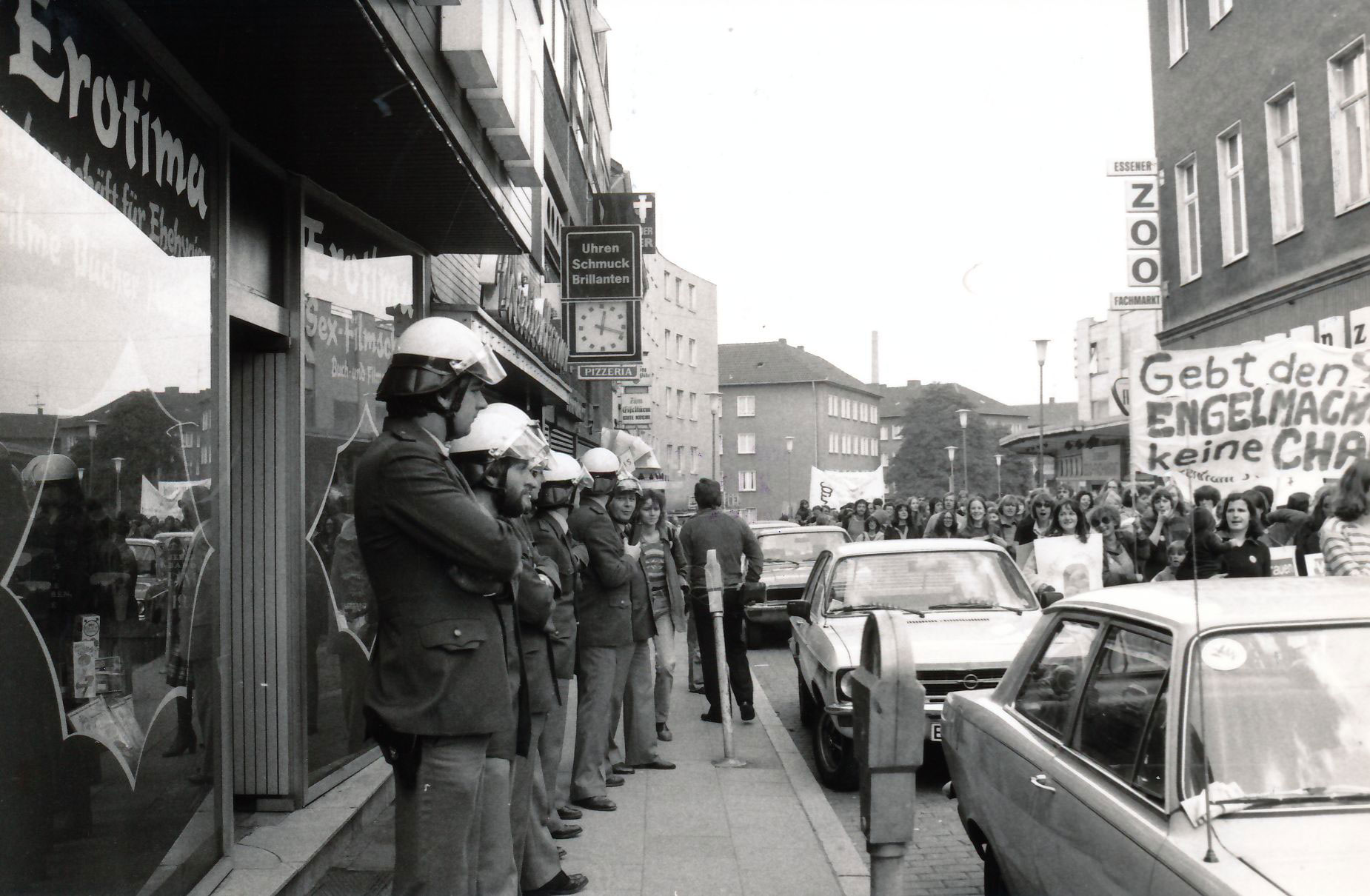 Demonstrantinnen ziehen mit dem Transparent mit der Aufschrift „Gebt den Engelmachern keine Chance“ durch eine schmale Straße in der Innenstadt; neun Polizisten stehen als Schutz vor einem Sexshop mit der Aufschrift “ Erotima. Fachgeschäft für Ehehygiene. Filme Bücher Magazine. Sex-Filmschau“; parkende Autos