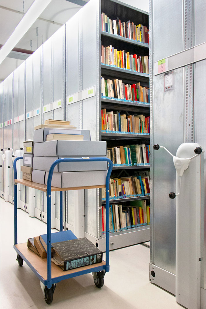 Archivwagen mit Kisten und Aktenordnern vor Rollregal mit Büchern im Magazin