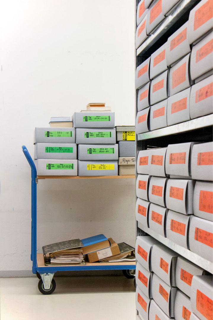 übereinander gestapelte Archivkisten im geöffneten Rollregal, Archivwagen mit Archivkisten und Aktenordnern