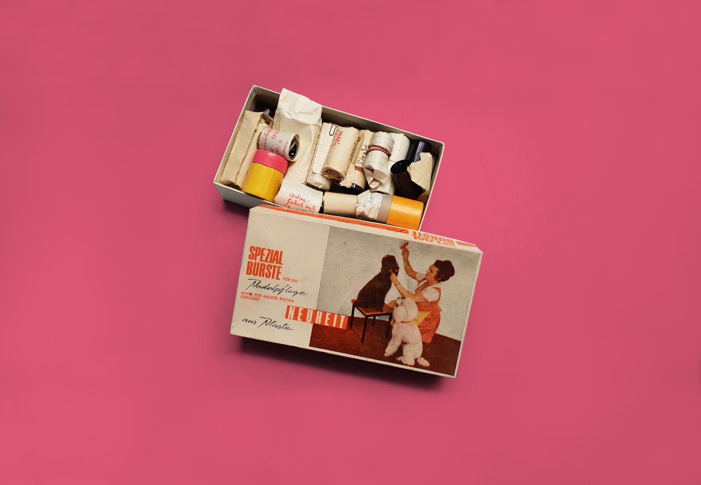 geöffnete Kiste mit Filmrollen; Deckel mit der Aufschrift „Spezialbürste für die Pudelpflege“ zeigt Abbildung einer Hundepflegerin bei der Arbeit mit zwei Pudeln