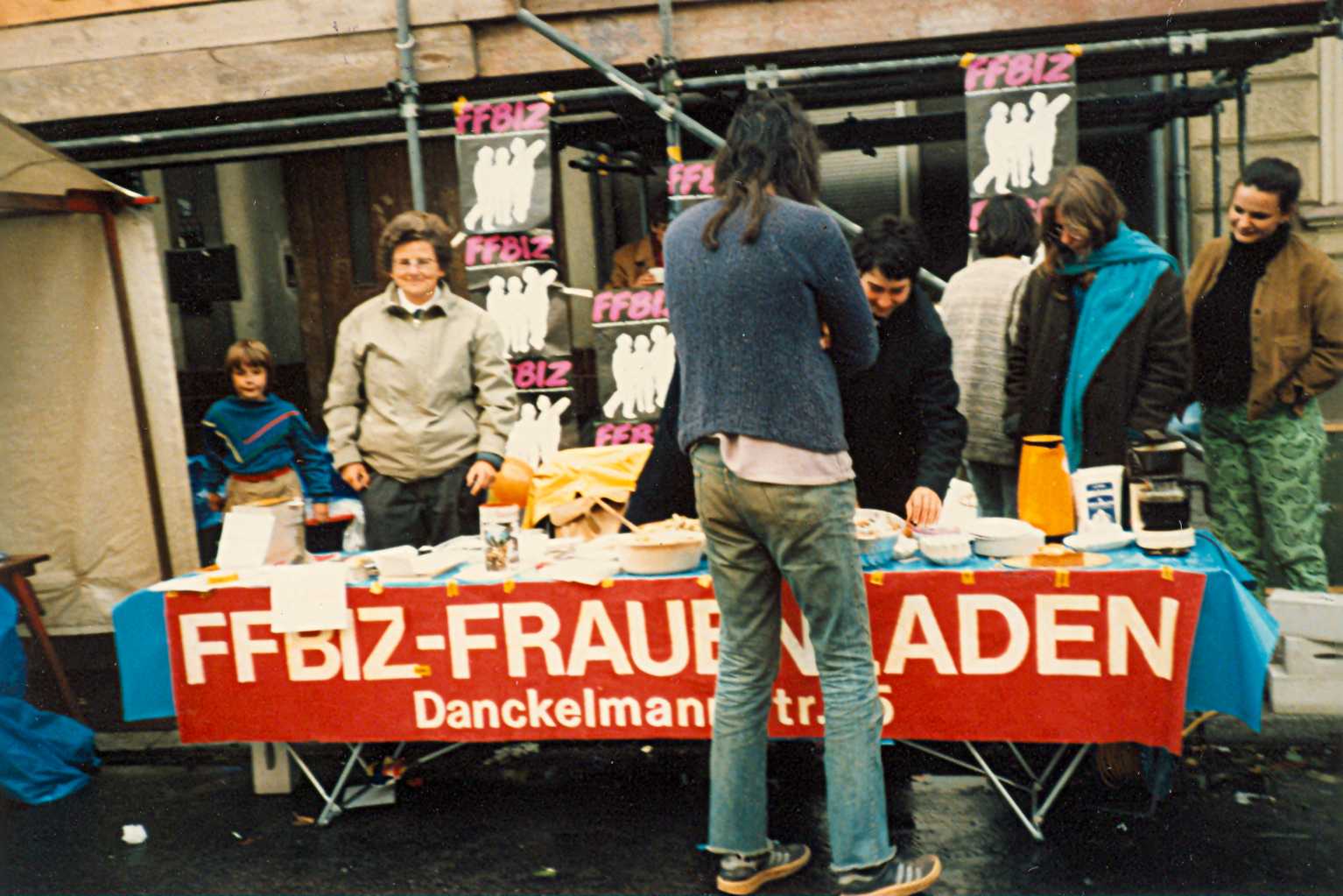 Gisela Vollradt steht neben anderen Frauen hinter einem Tisch mit Broschüren und anderen Informationsmaterialien auf der Straße vor einem Wohnhaus mit Baugerüst, an dem Info-Tisch hängt ein rotes Transparent bzw. Schild mit der Aufschrift FFBIZ-Frauenladen Danckelmannstraße 15, eine interessierte Frau steht vor dem Stand und betrachtet die ausgelegten Materialien; auf der rechten Tischseite Salat-, Kuchen- und Kaffeeausschank bzw. -verkauf; im Hintergrund FFBIZ-Plakate an den Stangen des Baugerüstes