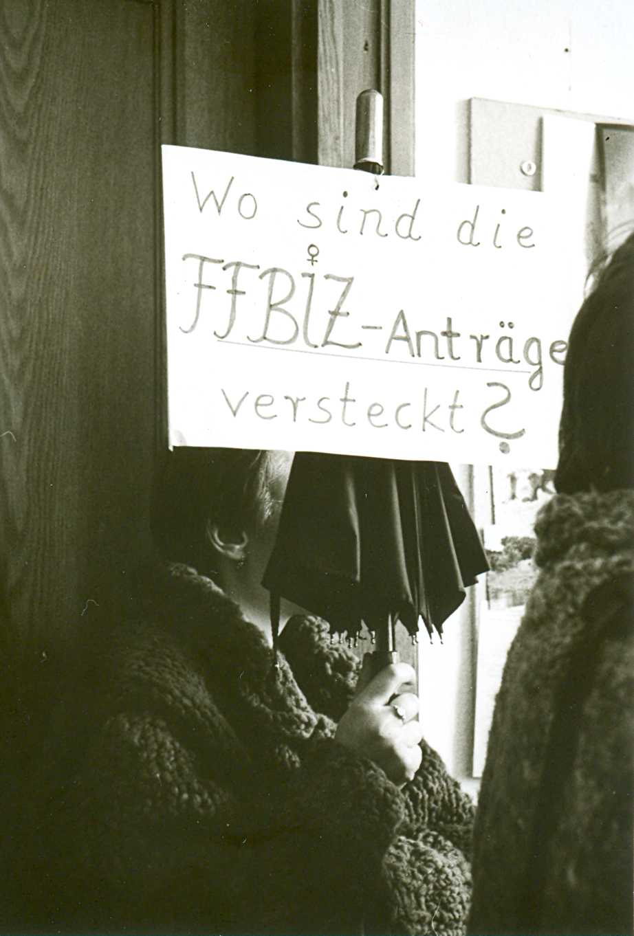 Ulrike Marski steht vor der Tür und hält einen Regenschirm hoch, an dem ein Protest-Plakat mit der Aufschrift „Wo sind die FFBIZ-Anträge versteckt?“ festgemacht ist; neben ihr protestiert eine weitere FFBIZ-Frau in den Büroräumen der Senatsverwaltung