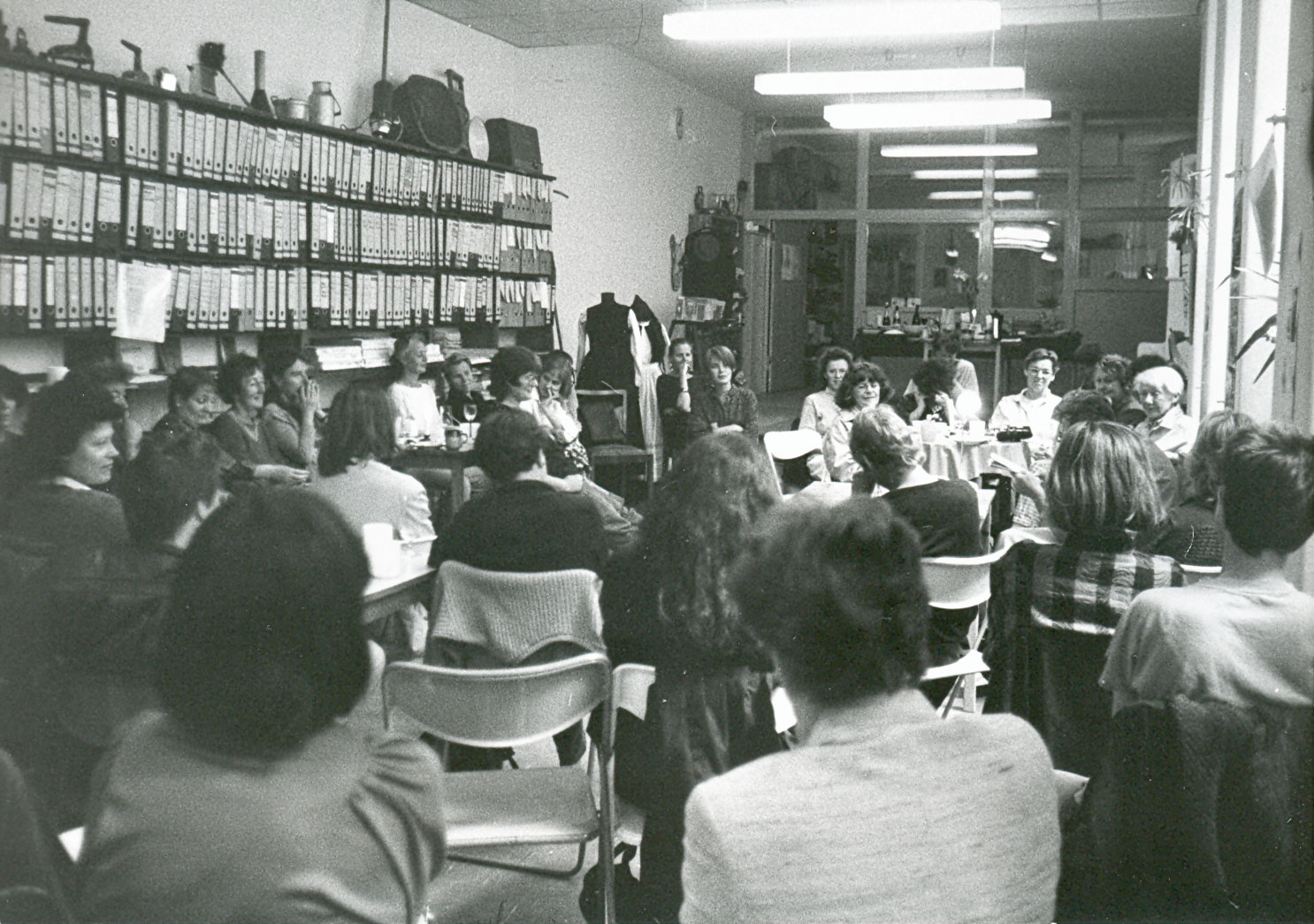 Zum Teil Rückenansicht: Gruppe von ca. dreißig Besucherinnen und Mitarbeiterinnen sitzt im Halbkreis im Veranstaltungsraum des FFBIZ um Berta Waterstradt und hört ihrer Lesung zu; im linken Bildausschnitt Regalwand mit Akten und Büchern