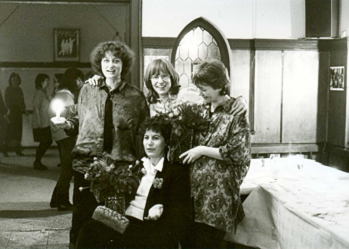 Gruppenfoto der FFBIZ-Mitfrauen Hilde Fauland-Weckmann, Sabine Spiesmacher, Ursula Nienhaus und Brigitte Pittelkau, zwei von ihnen halten Blumensträuße in Vasen, Hilde Fauland-Weckmann trägt eine Leuchte in der Hand; Ursula Nienhaus steht hinter ihnen, beide Arme um Hilde Fauland-Weckmann und  Brigitte Pittelkau gelegt; im Hintergrund Blick auf weiteren Raum der Gaststätte mit tanzenden Frauen